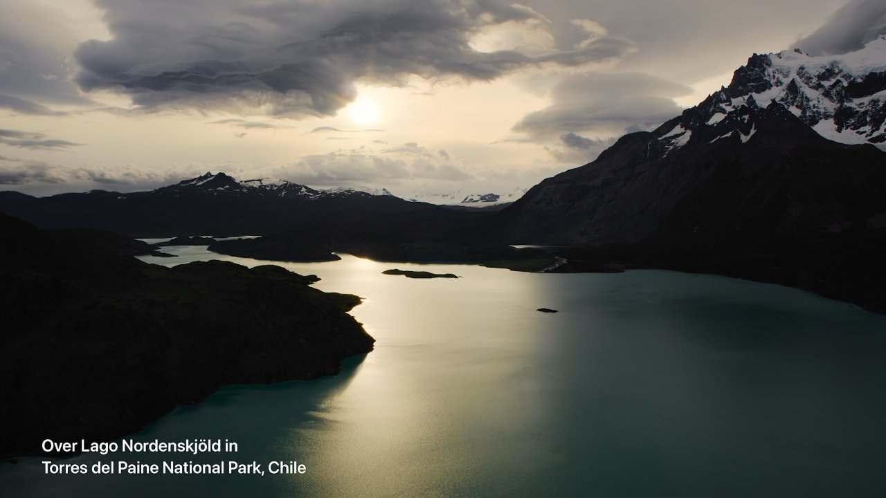 Sunset over a serene lake surrounded by mountains and dramatic clouds, reflecting soft light on the water surface.