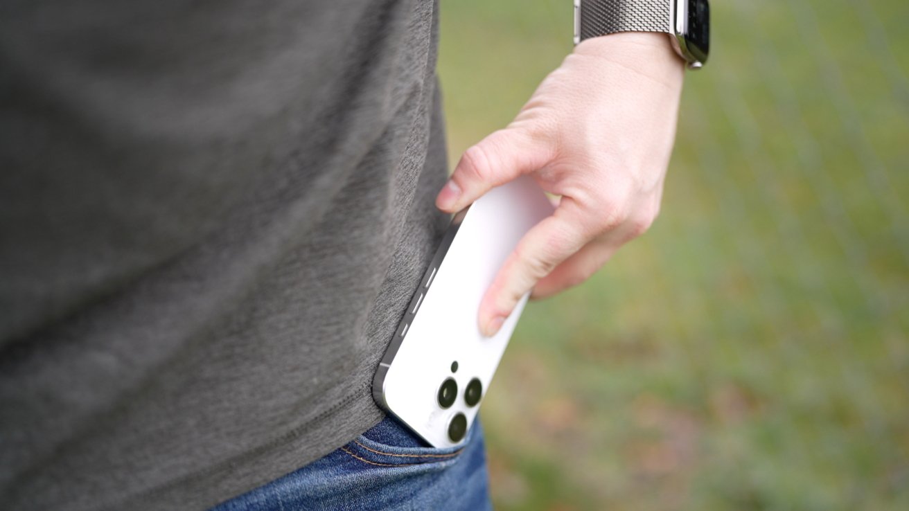 Person wearing a gray shirt and jeans placing a smartphone with three cameras into a pocket while wearing a metal watch.