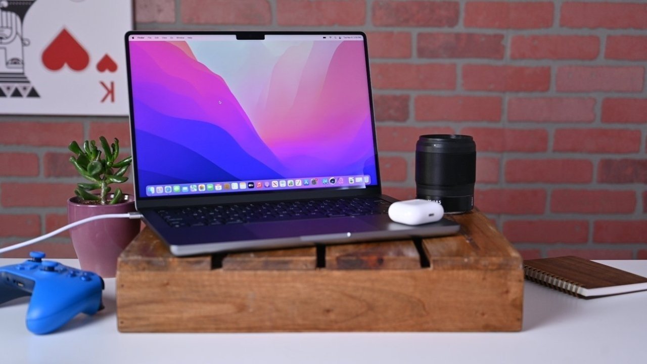 Laptop with colorful screen on a wooden stand, surrounded by a potted plant, camera lens, earbuds, blue game controller, and notebook against a brick wall.