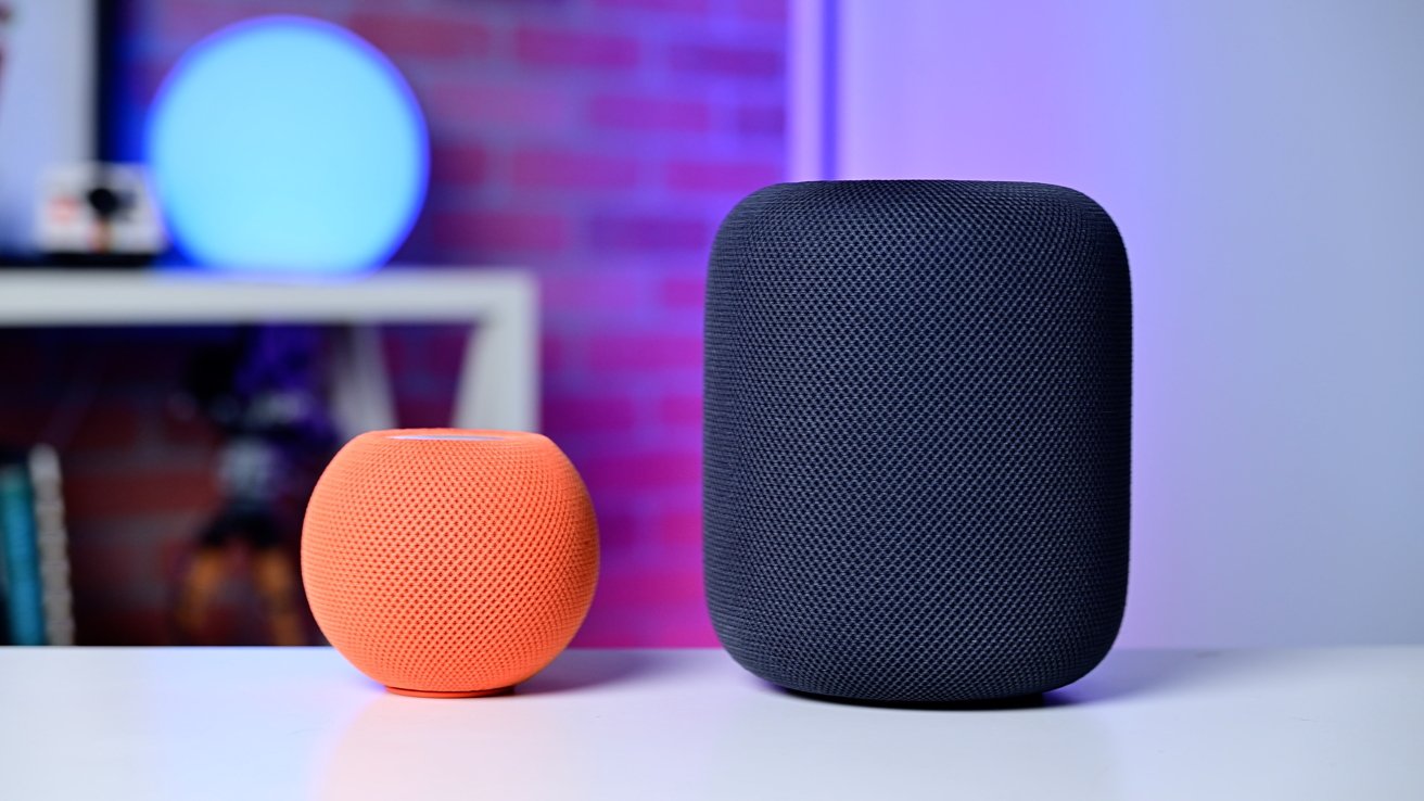 Two smart speakers, one small and orange, the other large and black, on a white surface with a blurred colorful background.