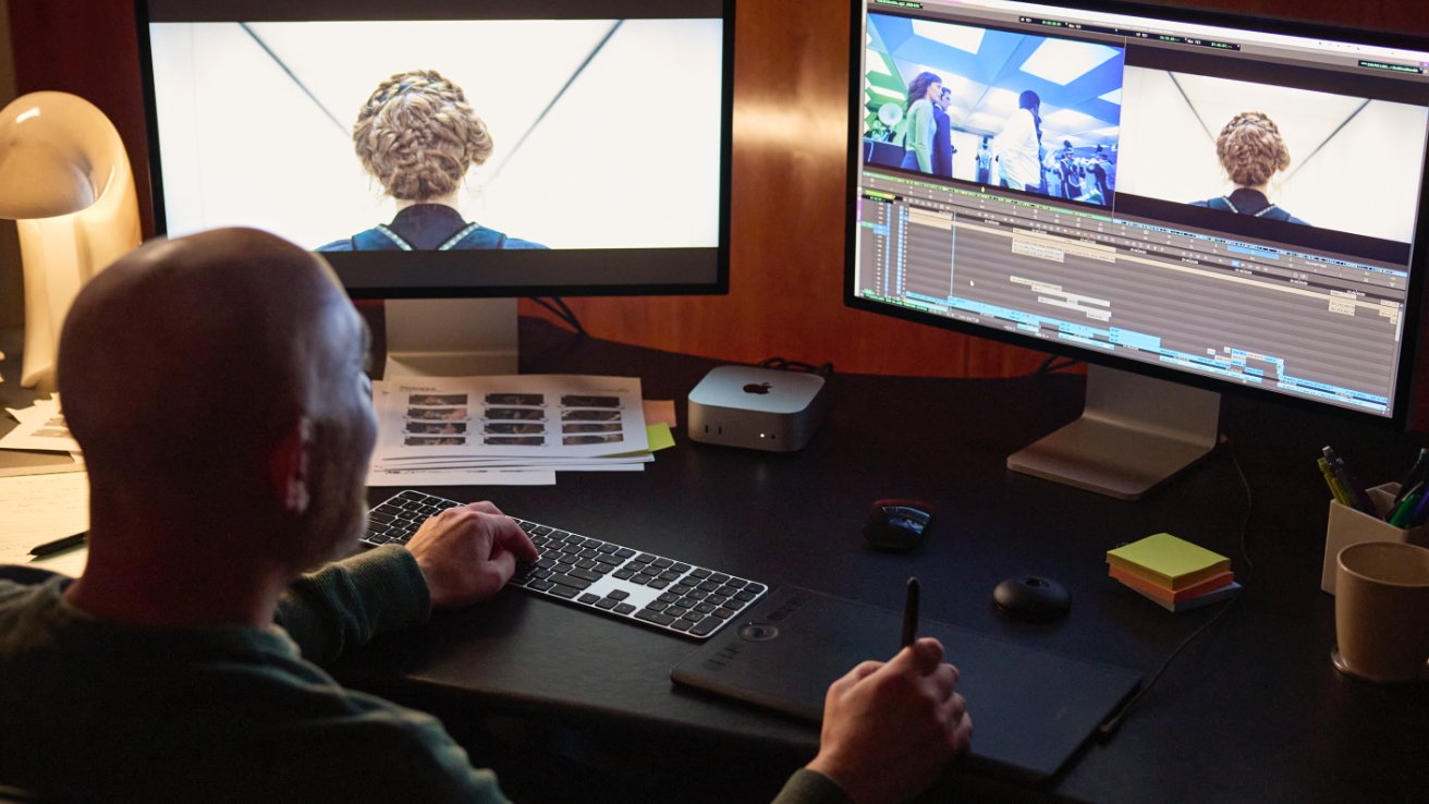Person editing video on dual monitors, using tablet and keyboard, surrounded by workspace items like lamp, papers, sticky notes, and a cup.