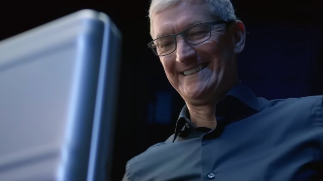 Smiling person with glasses, wearing a dark shirt, looking at a large, glowing screen in a dimly lit environment.