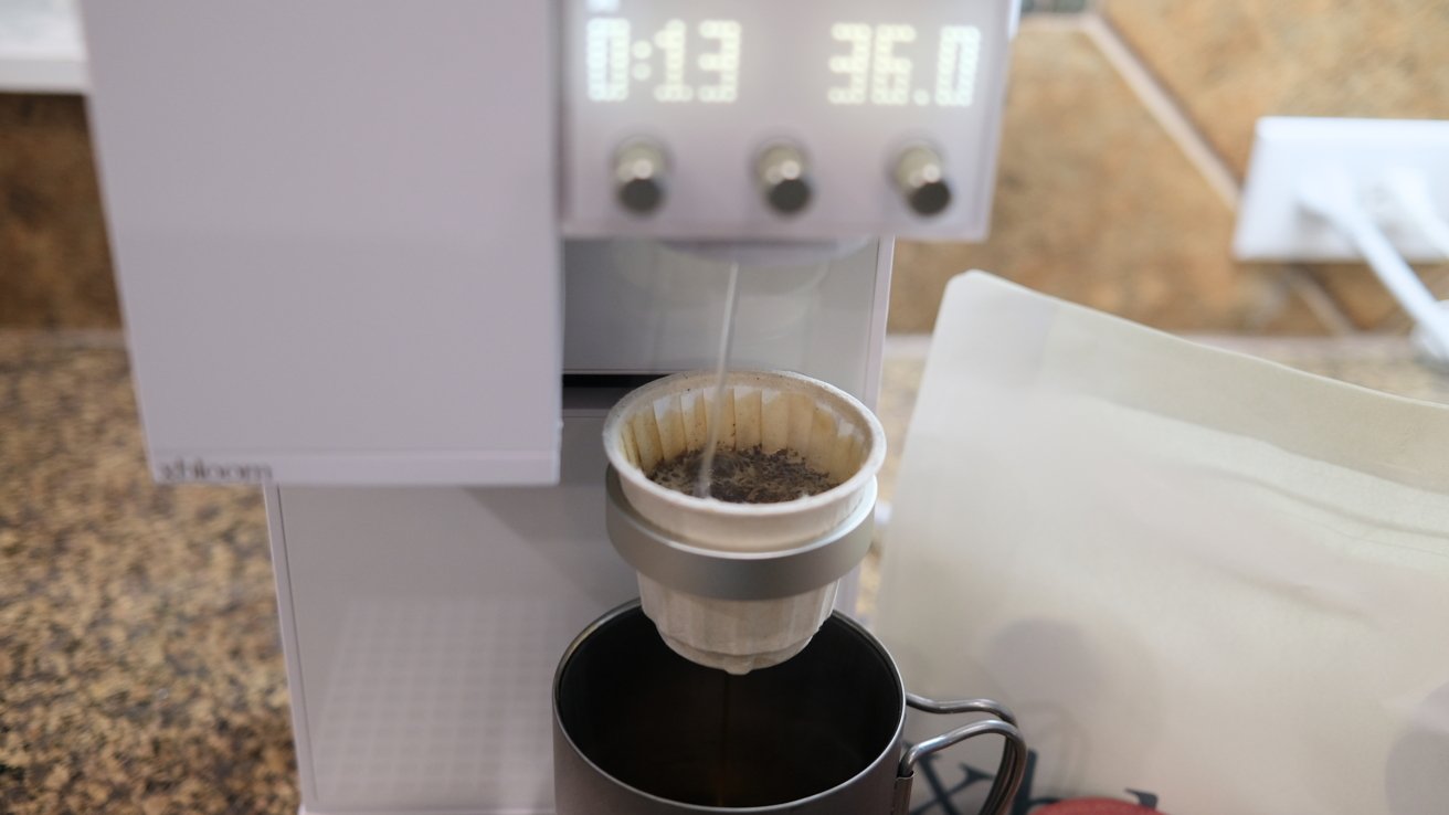 A white coffee maker dripping hot water through a white filter into a metal cup, with digital display showing time and temperature.