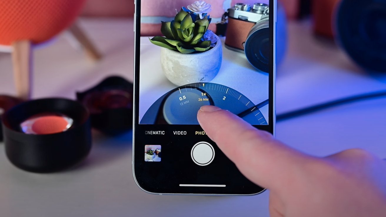Hand holds a smartphone capturing a photo of a plant in a pot beside a vintage camera, with focus slider displayed on screen.